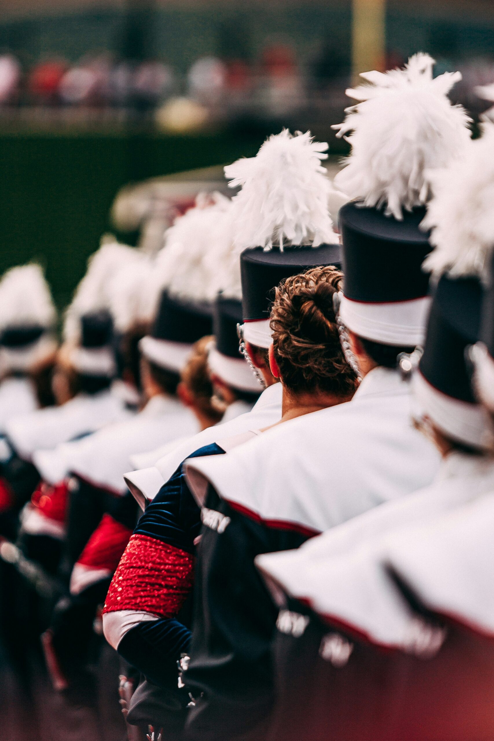 Marching band students in white uniforms and plumed shakos standing together in the stadium stands during a football game.